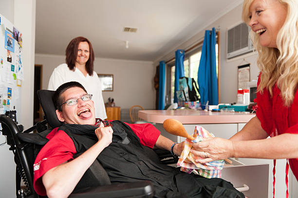 Man in wheelchair being assisted to wipe dry a spoon Young man being assisted to wipe dry a wooden spoon. disabled adult stock pictures, royalty-free photos & images