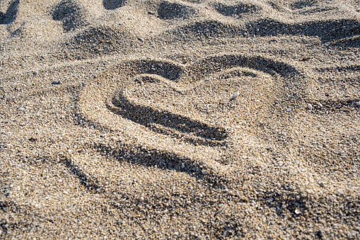 Stock photo showing heart shape drawn on sunny beach in sand with stick in soft, golden, wet sand on seaside coastline.