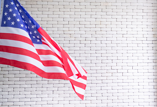 Close-up of the American flag is on the left side against a white brick wall background. 4th of July. Celebrate American National Day. Labor Day. Independence Day. Memorial Day.