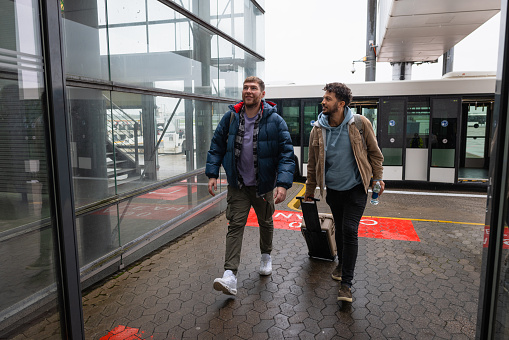 LGBTQI+ couple walking through an airport together ready to go on holiday. They are traveling to Germany for their vacation. They have just gotten off an airport shuttle bus.