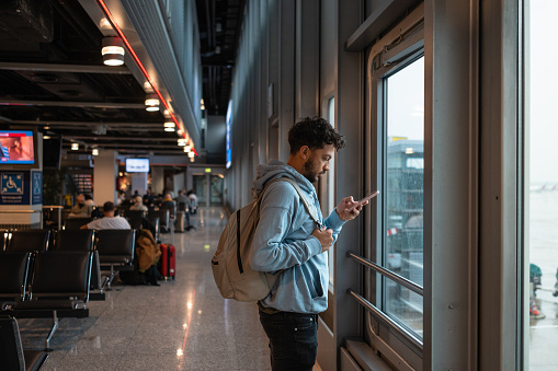 Side view of an LGBTQI+ man waiting in an an airport, going on vacation in Germany. He is standing by a window using his mobile phone.