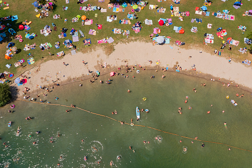 A beach with lots of people sunbathing and swimming in a lake in a summer sunday, aerial view.