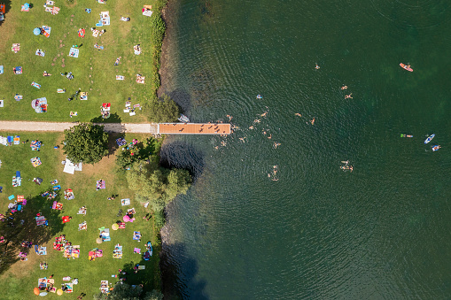 A grassy lake shore with a platform, lots of people sunbathing and swimming in a lake viewed from above in a summer sunday.