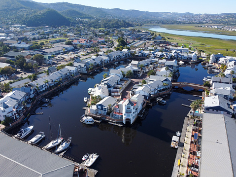 Aerial shot of the Knysna quays with a view of multiple multi coloured houses on the brown river with small private docks with boats in it and a small arched bridge connecting the island with the mainland