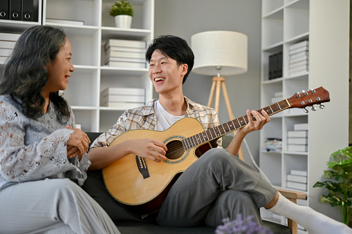 A happy young Asian grandson is singing and playing guitar to his grandmother, sitting on a sofa in the living room, and enjoying fun time at home together.