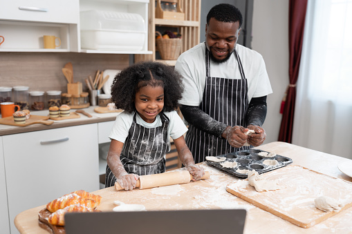 Happy African American kid girl learning cooking break or bakery with notebook computer at kitchen at home
