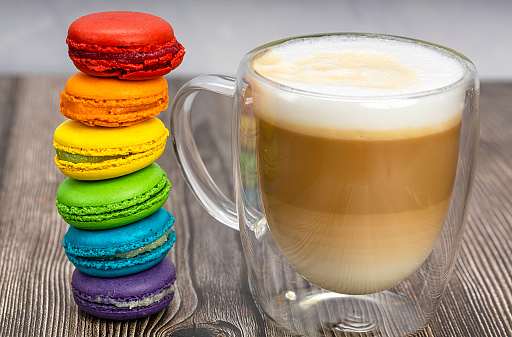 Multicolored macaroons in the colors of the LGBT rainbow flag and a transparent mug of cappuccino. The concept of freedom and love. Romance and sweets. Food photo.