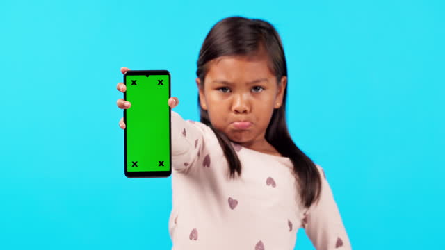 Phone, green screen and an unhappy girl on a blue background in studio for marketing or advertising. Kids, branding and a moody child holding chromakey mockup with tracking markers on a blank display