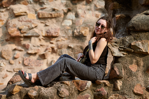 Attractive east asian woman in black sunglasses and black clothes sitting on ancient stone wall of old indian architecture buildings in Qutb Minar complex, candid young girl photo session in India