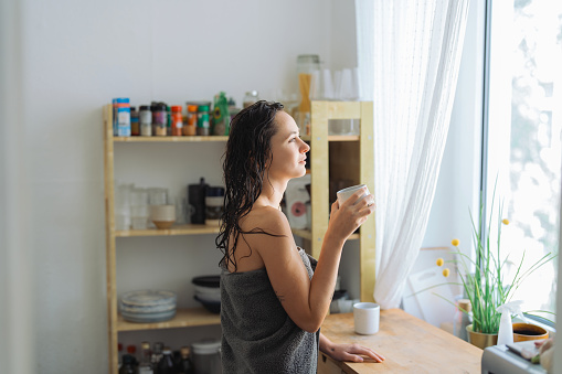 Woman after shower wearing a towel is drinking coffee in the morning on her kitchen and looking through the window