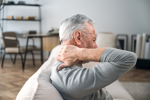 Elderly man rubbing hard pain in neck and massaging tense muscles. Feeling unwell, suffering from ache. Incorrect posture. Close up over shoulder view