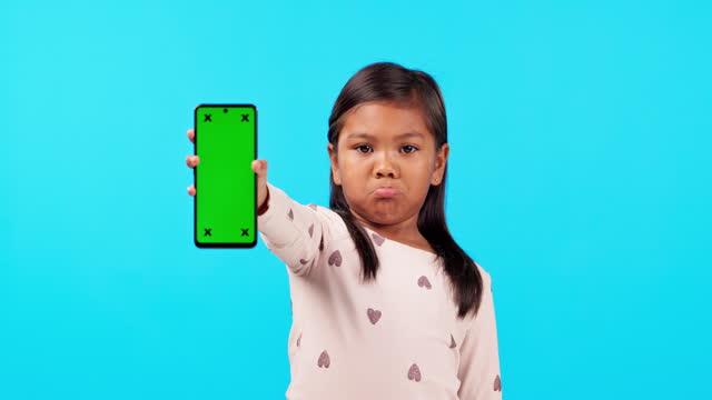 Phone, green screen and a moody girl on a blue background in studio for marketing or advertising. Kids, branding and an unhappy child holding chromakey mockup with tracking markers on a blank display
