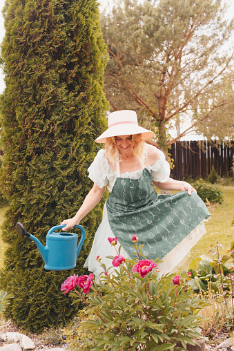 A beautiful mature girl with short blonde blonde hair in a blue vintage fashionable dress with a dark apron waters flowers from a large blue watering can and takes care of them in her beautiful summer garden in the backyard of her house in the village