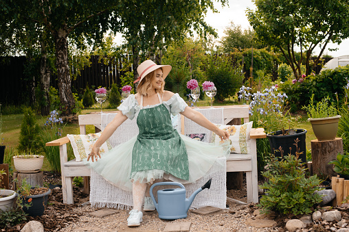 A mature beautiful woman in a beautiful fashionable hat with wavy short hair in a beautiful blue dress with an apron sits and rests in her cozy corner in the backyard on the lawn after taking care of her garden on a hot summer day in the village in summer