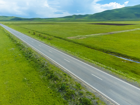 Drone view of an empty paved road in green meadows, mountains on a sunny day. Beautiful landscape with a roadway, sunlight, trees, green grass, clouds. Armenia