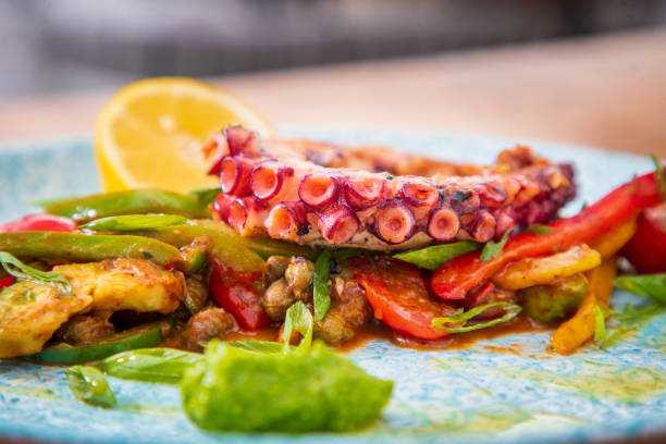 Close-up on juicy serving of grilled octopus in plate with pepper slices and avocado, lemon slice garnish, fine dining Mediterranean style