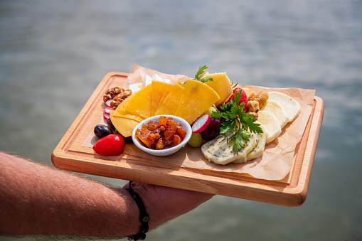 Carrying delicious assortment of cheese served on wax paper on wooden board with olives and nuts, sweet candied fruit cream in a bowl, outdoor fine dining appetizer