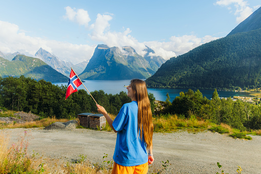Rear View of a female with long hair contemplating summer with a flag of Norway, with a background view of beautiful fjord during the summertime