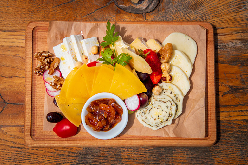Directly above delicious assortment of cheese on wax paper served on wooden board with fresh fruit, olives and nuts, sweet candied fruit cream in a bowl, fine dining appetizer