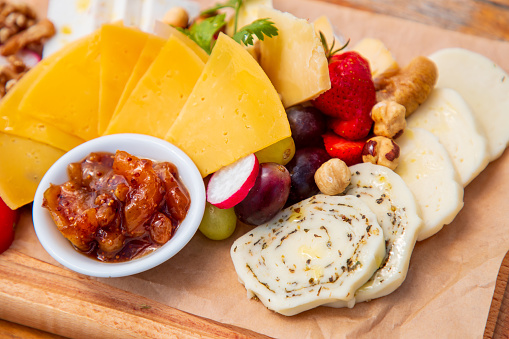 Delicious assortment of cheese on wax paper served on wooden board with fresh fruit, olives and nuts, sweet candied fruit cream in a bowl, fine dining appetizer