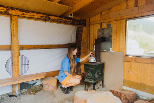 Side View of a female with long hair in a blue t-shirt contemplating a summer day in traditional Norwegian gapahuk, warming tea on old wooden oven in Scandinavia
