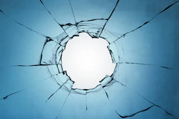 Broken glass with a hole on a blue background and with a light in the center. Texture of cracks on a broken window from explosions and gunshots