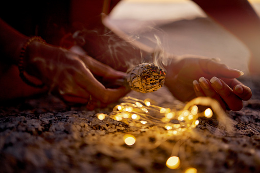 Hands, light and sage smoke at beach for healing or aromatherapy zen meditation. Woman, wellness and mental health in nature for mindfulness or spiritual chakra energy with fairy lights on ocean sand