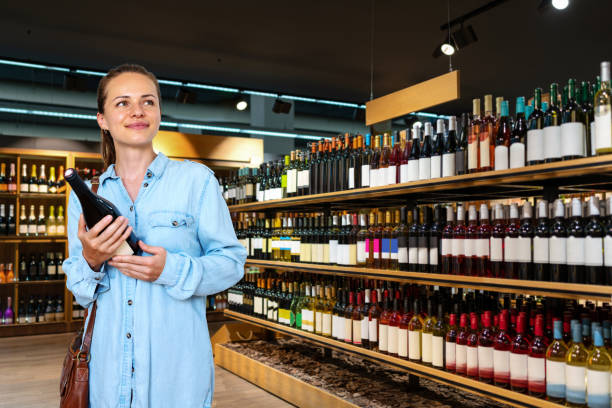 young adult brunette woman choosing wine in wine store and smiling. - wine bottle liquor store wine variation imagens e fotografias de stock