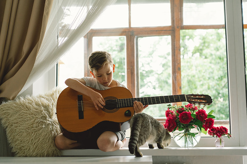 Cute boy learns to play the classical guitar on the windowsill near the window with cat. Cozy home. Summer holidays lifestyle.