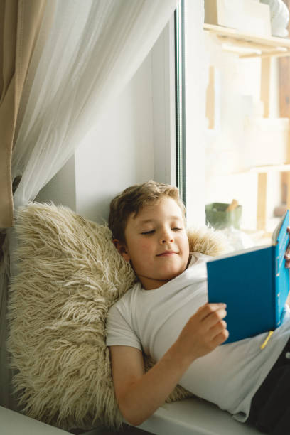 istock Cute boy reading a book on the windowsill near the window. 1533512457