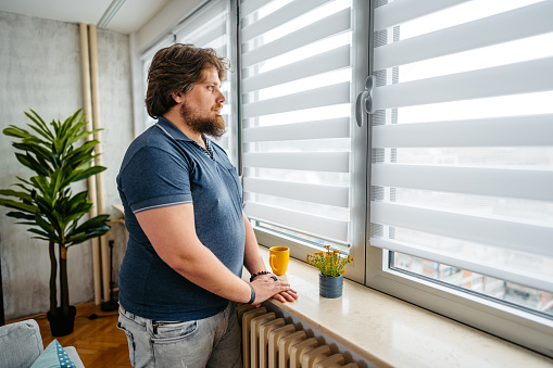 Portrait of a handsome young overweight man looking out the window of his apartment.