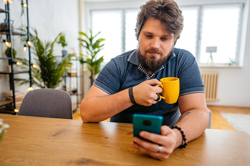 Smiling young overweight man relaxing at home, sitting at the kitchen table and texting on smartphone.