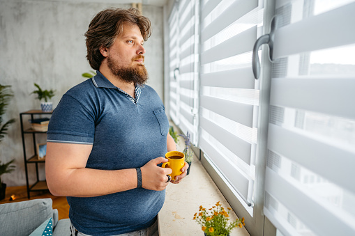 Overweight young man standing by the window at home and drinking coffee.