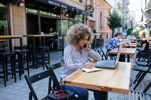 Young business focused woman is sitting in cafe and working on laptop. She is talking on the mobile phone and writing a notes. Business on the go.