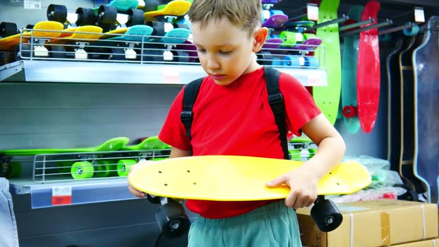 A cute boy holds a skateboard in his hands and examines it carefully in a sports department