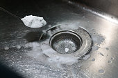 From a high angle view, a woman's hand is adding a mixture of sodium bicarbonate, vinegar, and hot water into a kitchen sink, down the drain.