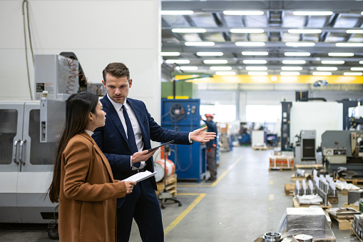 The manager debates with the customer about the actual order of products in their workshop. A white man and an Asian woman are looking at the production. The white man is in a blue business suit with a tie, the visitor is in a coat. Bright manufacturing plant with metalworking machinery. Business meeting, copy space, bright,