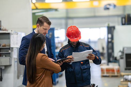 A white manager in a business suit and an Asian colleague in a coat are discussing the distribution of goods with a worker in a worker's suit and wearing a helmet in a warehouse area. They look at delivery vans and warehouse racks.
