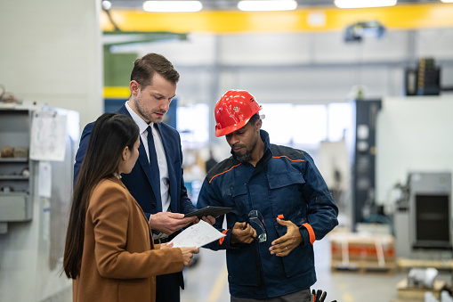 A white manager in a business suit and an Asian colleague in a coat are discussing the distribution of goods with a worker in a worker's suit and wearing a helmet in a warehouse area. They look at delivery vans and warehouse racks.