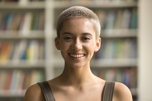 Cheerful beautiful orthodontist patient, teenage girl with teeth braces looking at camera, smiling. Happy pretty young high school, college teen student front portrait in campus library