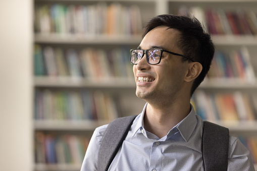 Happy nerdy teenager student guy in eye glasses and backpack standing in library, looking away, thinking over higher college education, homework task, knowledge. Candid portrait
