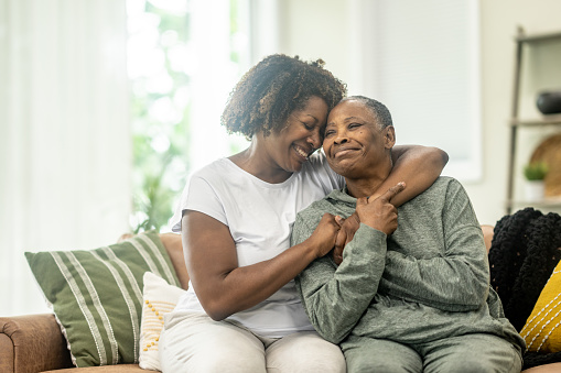 A middle aged woman of African decent wraps her arm around her Mother as she embraces her and they pose for a portrait.  They are both dressed comfortably and are laughing as they enjoy their time together.