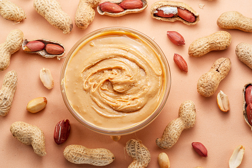 Bowl of peanut butter and peanuts around it on beige background. Top view.