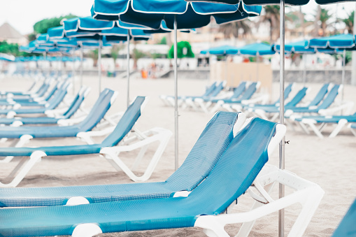 Perspective view on a row of blue empty sunbeds and beach umbrellas in a summer cloudy day