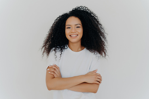 Pleasant young woman with curly hair, hands crossed, white t-shirt, broad smile, shy expression, indoor pose, natural beauty. Expressive portrait.