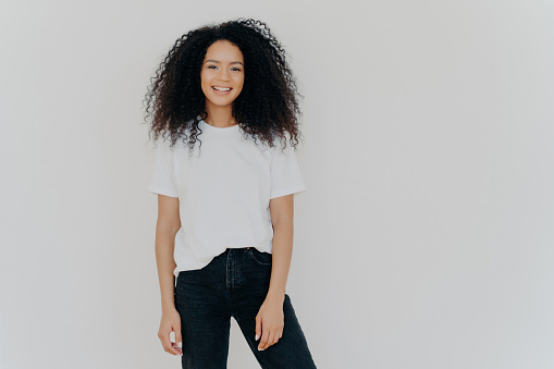 Smiling woman with curly hair, slim figure, white t-shirt, black jeans, joyful expression, models on white background. Copy space for ads.