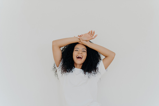 Overjoyed curly-haired lady, hands on forehead, laughing with mouth wide open, energetic vibes. Casual attire, white studio backdrop. Hilarious joke!