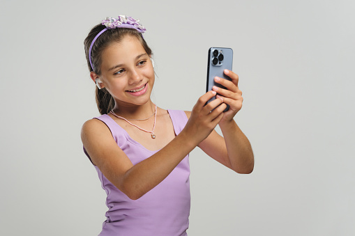 Studio shot portrait of a beautiful child girl using smartphone and wireless earphones, selfie concept