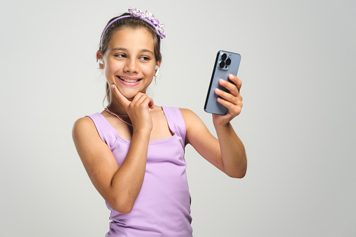 Studio shot portrait of a beautiful child girl using smartphone and wireless earphones, selfie concept