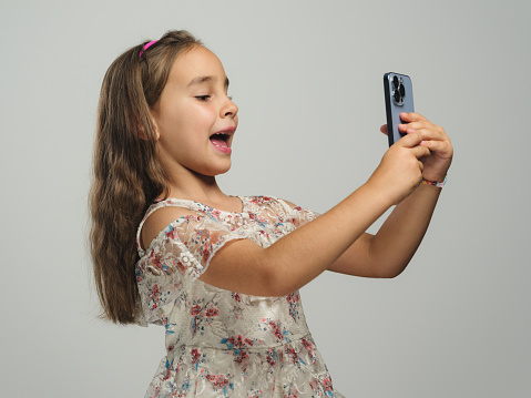 Studio shot portrait of a beautiful child girl using smartphone and wireless earphones, selfie concept